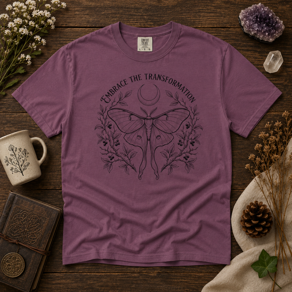 Berry color t-shirt with luna moth design and text 'Embrace the Transformation' on a wooden surface with decorative items.